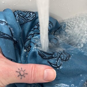 Pode incluir: Um tecido azul com um padr&atilde;o floral preto est&aacute; sendo lavado em &aacute;gua corrente. O tecido est&aacute; submerso em uma pia com bolhas se formando. Um dedo com uma tatuagem de sol &eacute; vis&iacute;vel.