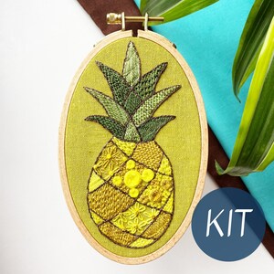May include: Embroidered pineapple design in a wooden embroidery hoop. The pineapple is yellow and green with a variety of stitches. The hoop is on a blue and brown surface. The word "KIT" is written in white on a blue circle.