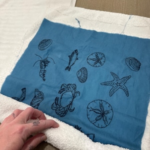Pode incluir: Um tecido quadrado azul com desenhos a tra&ccedil;o preto de criaturas marinhas, incluindo um caranguejo, uma estrela do mar e um peixe, &eacute; contornado por um tecido de felpa branco. Provavelmente faz parte de uma toalha ou de um artigo decorativo com tema de praia.