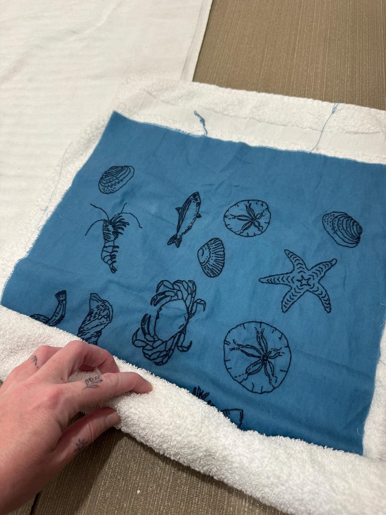 Pode incluir: Um tecido quadrado azul com desenhos a tra&ccedil;o preto de criaturas marinhas, incluindo um caranguejo, uma estrela do mar e um peixe, &eacute; contornado por um tecido de felpa branco. Provavelmente faz parte de uma toalha ou de um artigo decorativo com tema de praia.