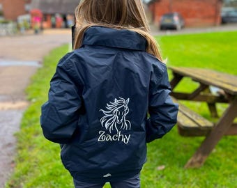 May include: A navy blue jacket featuring a white embroidered graphic of a horse head with flowing mane and the name Zoachy written in cursive script below the design. The jacket has a high collar and long sleeves, worn over dark trousers.