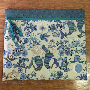 May include: A white zippered pouch with a teal blue and white wave pattern on the top. The pouch features a colourful design with mermaids, flowers, and skulls. The text "The day before This day" is printed on the bottom.