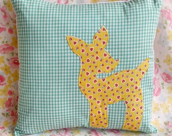Op de afbeelding: Een groen en wit gingham kussen met een gele herten applique. Het hert is gemaakt van een gele stof met een rood en geel bloemenpatroon.