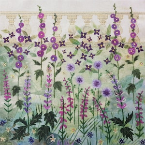 May include: A close-up of an embroidered wall hanging featuring a garden scene with purple, pink, and white flowers. The flowers are arranged in front of a white picket fence.