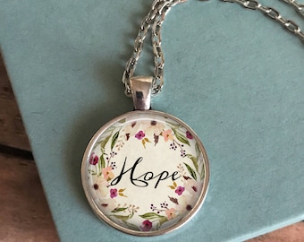 May include: Silver-toned pendant necklace featuring a round pendant with the word "Hope" in elegant script. The pendant is encircled by a floral wreath in shades of pink, purple, and green. The necklace rests on a light blue surface.