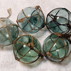 May include: Five spherical glass fishing floats in a light blue-green hue, each encased in a hand-knotted brown or tan rope netting. The glass surfaces are translucent and smooth, featuring small air bubbles trapped within the material structure.