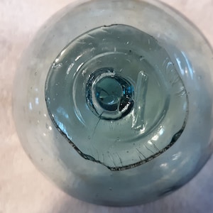 May include: A round, pale blue glass fishing float featuring a thick, circular glass seal at the center. The surface of the translucent glass sphere displays small air bubbles and a distinct, textured pontil mark where the glass was gathered.