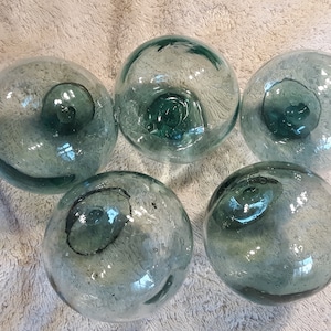May include: A collection of five spherical glass floats, each featuring a translucent pale blue-green tint. These round objects display visible air bubbles and a distinct circular seal mark on their surface, characteristic of hand-blown glass.