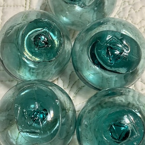 May include: Five clear, teal-colored glass floats with a central, recessed design. The floats are round with a slightly flattened top and bottom. The glass has a bubbly texture, and the central design appears to be a geometric shape.