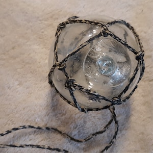 May include: A clear, spherical glass float encased in a decorative, knotted netting made of black and tan twisted cord. The netting features a repeating diamond pattern and includes two long, loose cord ends extending from the bottom of the sphere.