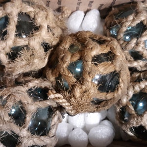 May include: Six glass floats wrapped in natural twine, with a blue-green tint, are packed in a box with white packing peanuts.
