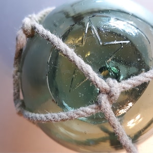 May include: A clear glass fishing float with a star design etched into the surface. The float is wrapped in a thick, gray rope. The float is a sphere, and the rope is knotted around it.