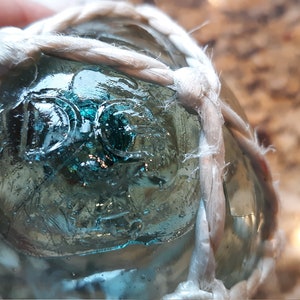 May include: Close-up of a blue glass fishing float with white rope netting. The float has the letters "D.C." embossed on the surface.