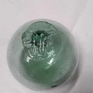 May include: A clear, round, glass float with a green tint. The top of the float has a textured, irregular opening. The float is likely used for decoration or as a fishing float.