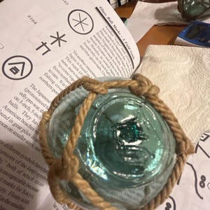 May include: A close-up of a light blue glass fishing float with a brown rope tied around it. The float has a mark on it that looks like a stylized "M".
