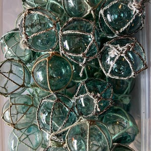 May include: A collection of green glass fishing floats, each encased in a white net. The floats are various sizes and shapes, and some have a slight blue tint.