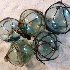 May include: Five glass fishing floats with a light blue tint and brown rope netting. The floats are arranged in a cluster on a white fuzzy surface.