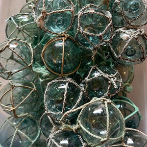 May include: A collection of green glass fishing floats, each encased in a woven net. The floats are various sizes and shapes, with some having a more rounded shape and others being more elongated. The nets are a light brown color and are woven in a tight pattern.