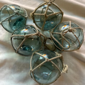 May include: A collection of six clear glass fishing floats with a blue-green hue. Each float is encased in a knotted, natural-colored rope netting. The floats are arranged on a white fabric surface.
