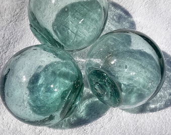 May include: Three round, green glass floats with a textured surface. The floats are on a white, fluffy surface.