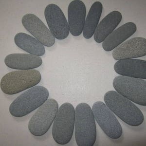 May include: A collection of eighteen smooth, oval-shaped gray stones arranged in a circular pattern. Each stone features a uniform, matte texture and varies slightly in shade from light gray to dark charcoal, creating a balanced, radial display.