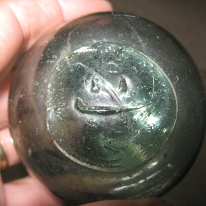 May include: A round, green glass fishing float with a circular design etched into the surface. The design appears to be a stylized face with two eyes and a mouth.