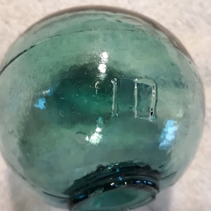 May include: A round, teal-colored glass float with a textured surface. The float has a visible seam and a rectangular marking. This decorative item is often used in nautical-themed decor.