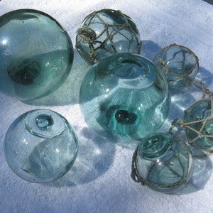 May include: Collection of vintage glass fishing floats in varying sizes. The floats are a translucent teal color, some encased in netting. The glass spheres have a classic nautical aesthetic, perfect for coastal decor or craft projects.
