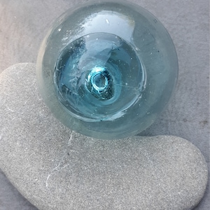 May include: A light blue glass sphere with a clear center, resting on a gray stone. The sphere has a smooth, rounded surface and a slightly textured appearance.