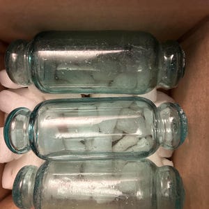 May include: Three aqua blue glass bottle stoppers with rounded ends. The stoppers are sitting in a box with white packing peanuts.
