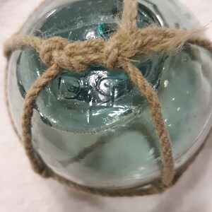 May include: A clear, round glass fishing float with a teal hue, wrapped with natural jute rope. The rope is knotted around the float, creating a decorative accent. The float has a vintage, nautical aesthetic.