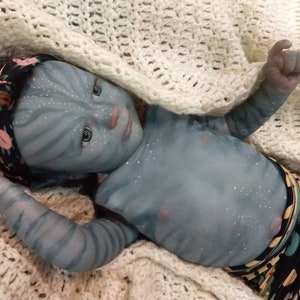 K&ouml;nnte beinhalten: Eine blaue Baby-Puppe mit einem schwarzen und rosa Stirnband, die auf einer wei&szlig;en H&auml;keldecke liegt. Die Puppe tr&auml;gt eine bunte gemusterte Windel.