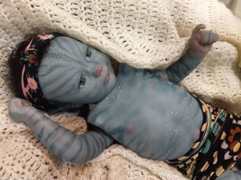 K&ouml;nnte beinhalten: Eine blaue Baby-Puppe mit einem schwarzen und rosa Stirnband, die auf einer wei&szlig;en H&auml;keldecke liegt. Die Puppe tr&auml;gt eine bunte gemusterte Windel.