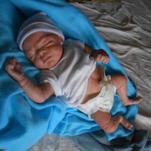 K&ouml;nnte beinhalten: Ein neugeborenes Baby in einem wei&szlig;en Strampler und einer rosa-wei&szlig;en M&uuml;tze schl&auml;ft auf einer blauen Decke. Das Baby hat eine kleine Nabelschnurklemme am Bauchnabel.