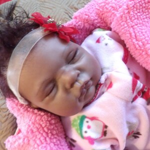 K&ouml;nnte beinhalten: Eine schlafende Babypuppe mit dunkelbraunen Haaren, die einen rosa-wei&szlig;en gemusterten Schlafanzug und ein wei&szlig;es Stirnband mit einer roten Blume tr&auml;gt.