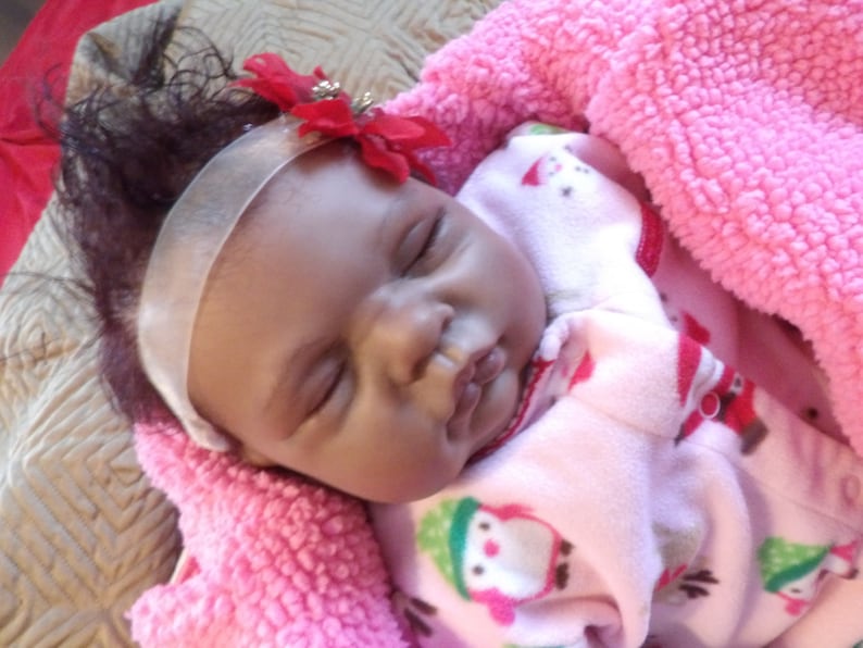 K&ouml;nnte beinhalten: Eine schlafende Babypuppe mit dunkelbraunen Haaren, die einen rosa-wei&szlig;en gemusterten Schlafanzug und ein wei&szlig;es Stirnband mit einer roten Blume tr&auml;gt.