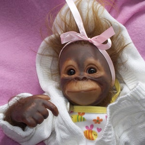 K&ouml;nnte beinhalten: Eine Pl&uuml;sch-Orang-Utan-Puppe, die einen wei&szlig;en Pullover mit einer rosa Schleife und ein gelbes und wei&szlig;es gemustertes Kleid tr&auml;gt.