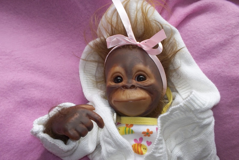 K&ouml;nnte beinhalten: Eine Pl&uuml;sch-Orang-Utan-Puppe, die einen wei&szlig;en Pullover mit einer rosa Schleife und ein gelbes und wei&szlig;es gemustertes Kleid tr&auml;gt.