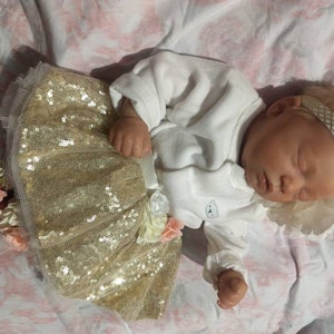 K&ouml;nnte beinhalten: Eine schlafende Babypuppe, die einen goldenen Paillettenrock mit einem wei&szlig;en Strampler und ein wei&szlig;es Stirnband mit goldener Verzierung tr&auml;gt.