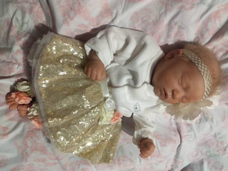 K&ouml;nnte beinhalten: Eine schlafende Babypuppe, die einen goldenen Paillettenrock mit einem wei&szlig;en Strampler und ein wei&szlig;es Stirnband mit goldener Verzierung tr&auml;gt.