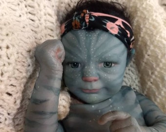 Könnte beinhalten: Eine blaue Baby-Puppe mit einem Stirnband, bemalt, um einem Charakter aus dem Film Avatar zu ähneln. Die Puppe hat einen hellblauen Körper mit dunkelblauen Markierungen und eine hellrosa Nase.