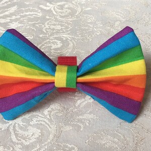 May include: A colorful rainbow-striped bow tie. The bow tie features a vibrant spectrum of colors, including red, orange, yellow, green, blue, and purple. The bow tie is made of fabric and has a small red and yellow center.