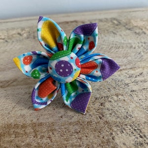 May include: A colorful fabric flower with a white background and a variety of colors including purple, blue, green, yellow, and orange. The flower has a polka dot pattern.