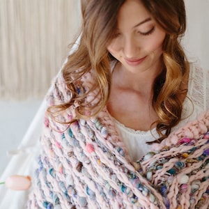 Może przedstawiać: Różowy i wielokolorowy, gruby pled z fakturowanym, tkanym wzorem. Pled jest zarzucony na ramiona osoby.