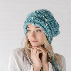 May include: A teal knitted hat with small white flower accents. The hat is made of a chunky yarn and has a slouchy fit. The hat is a stylish accessory for cold weather.