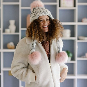 May include: A woman wearing a white faux fur jacket with a grey and pink knit hat with pom poms. The hat has long braids with pom poms at the ends.