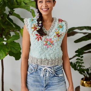 May include: A light blue knitted vest with a V-neck and floral embellishments. The vest has a grey trim at the bottom. The model is wearing denim shorts.