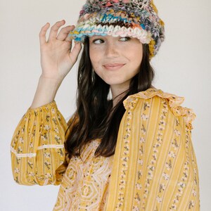 May include: A woman wearing a yellow and white floral patterned blouse with a multi-colored crocheted hat. The hat has a wide brim and is made of a variety of different colored yarns.