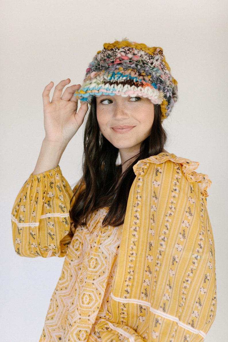 May include: A woman wearing a yellow and white floral patterned blouse with a multi-colored crocheted hat. The hat has a wide brim and is made of a variety of different colored yarns.