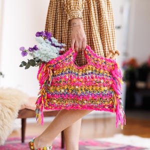 Puede incluir: Un bolso tote tejido a mano, vibrante, en tonos rosa, amarillo y azul, adornado con flecos. La bolsa está llena de flores y agujas de tejer. La persona lleva sandalias florales y un vestido de cuadros marrón.